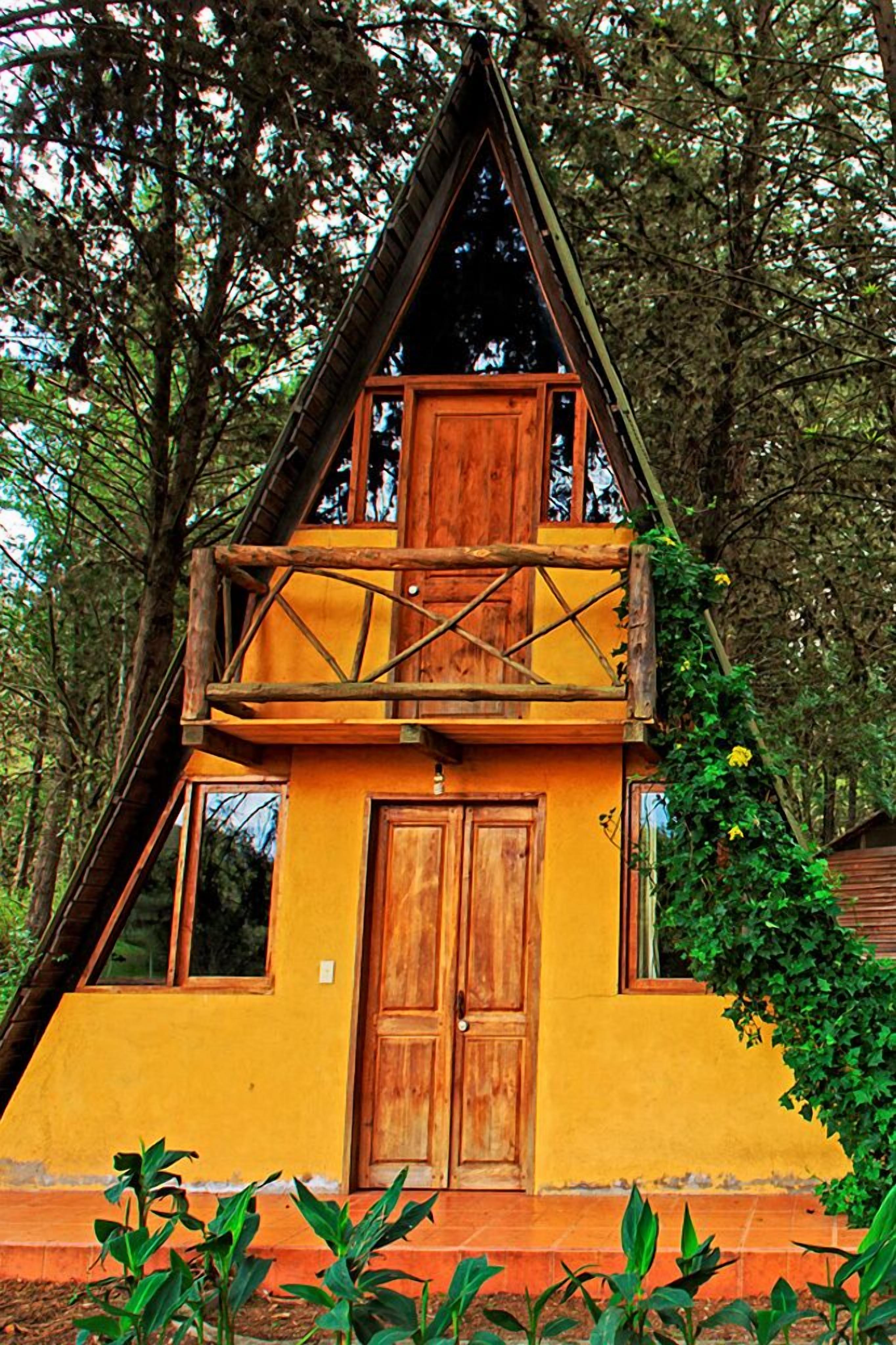 Una cabaña de madera de dos pisos con balcón, rodeada de árboles.