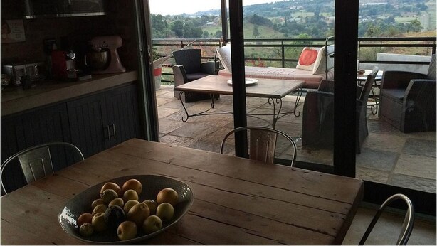 A dining area with a table and chairs, a bowl of fruit, and a view of the countryside through large windows. - The Warehouse (Krugersdorp)