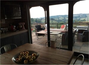 A dining area with a table and chairs, a bowl of fruit, and a view of the countryside through large windows. - The Warehouse (Krugersdorp)