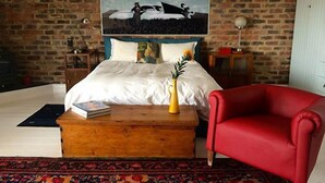 A bedroom with a brick wall, a bed with white bedding, a red armchair, a wooden nightstand, a bedside table with a lamp, and a poster on the wall. - The Warehouse (Krugersdorp)