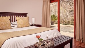 A hotel room with a large bed, a bedside table with a tray of toiletries, and a view of a desert landscape through a sliding glass door. - Riverwalk B and B (Clarens)
