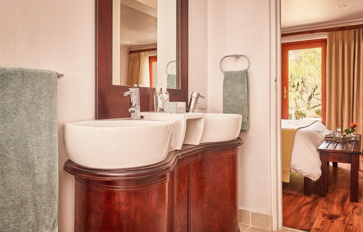 Une salle de bain avec deux lavabos, un miroir et une vue sur une chambre à coucher comprenant un lit et une table.