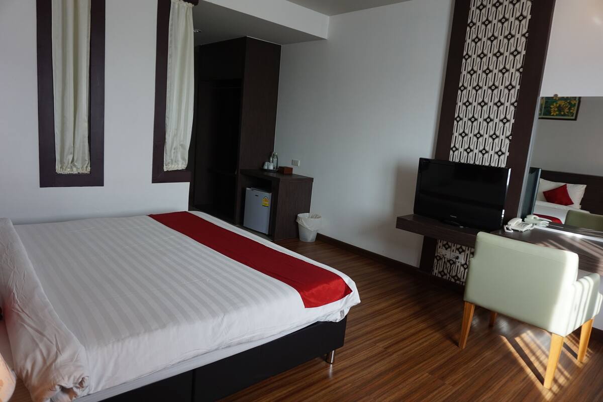 A hotel room with a bed, a desk with a TV, a chair, a small cabinet, and a patterned wall decoration.