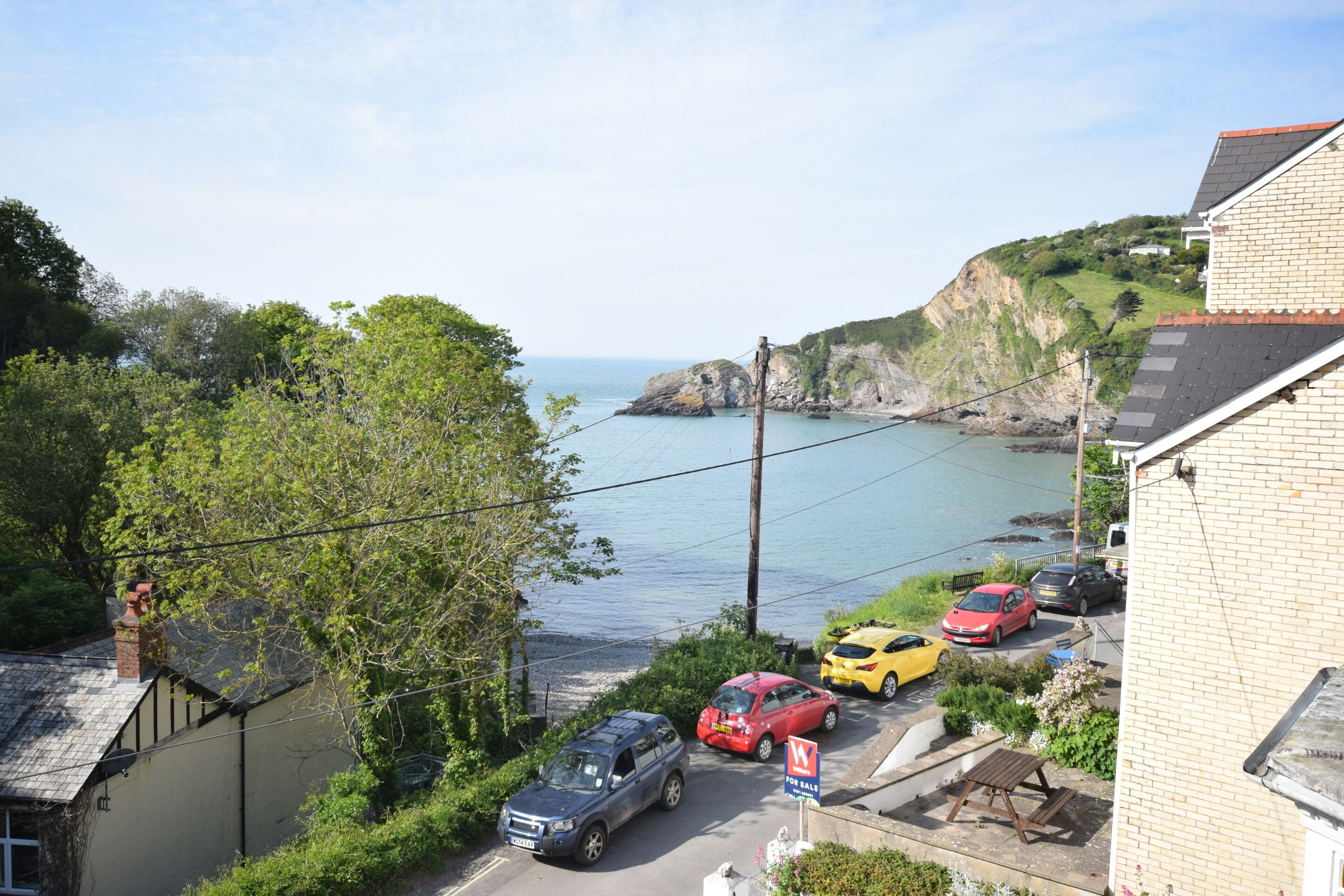 Un quartier résidentiel situé sur la côte, avec vue sur la mer, des voitures garées et une pancarte « À vendre ».