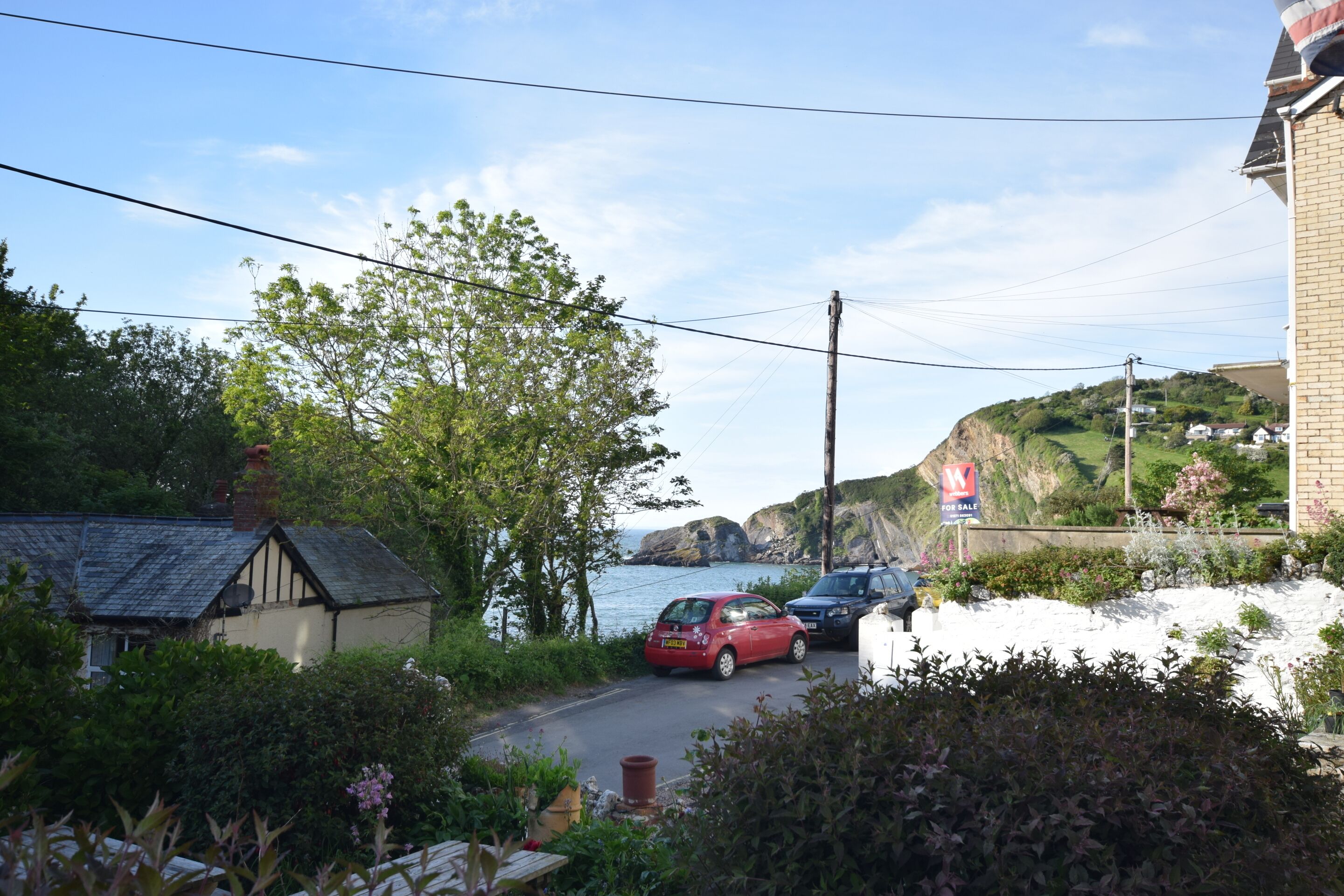 A coastal road with parked cars, a house, and a 'For Sale' sign.