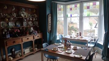 A dining area with a wooden table and chairs, a kitchen area with shelves of dishes and utensils, and a view through a bay window.
