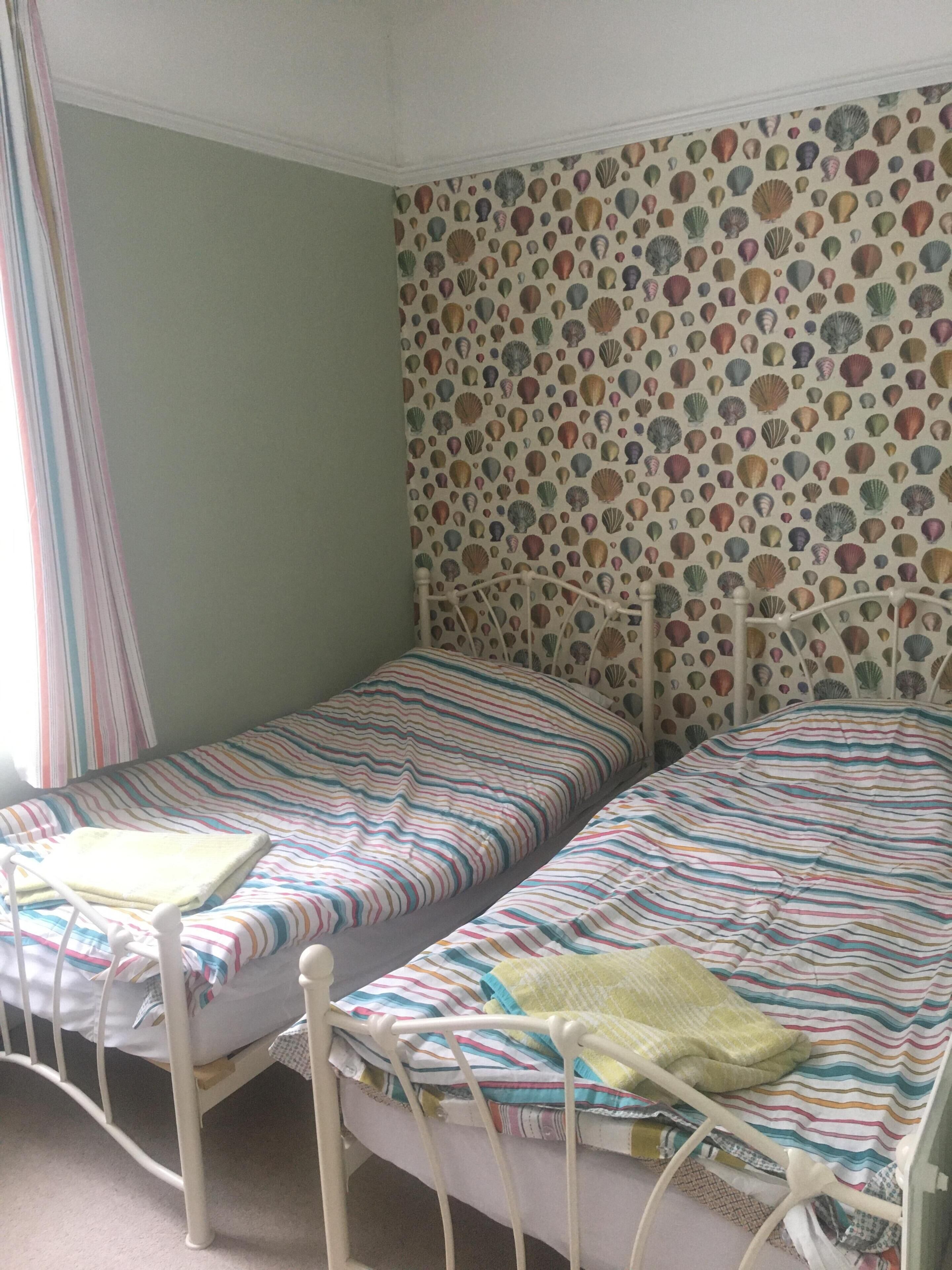 A room with two single beds, a patterned wallpaper, and a window with curtains.