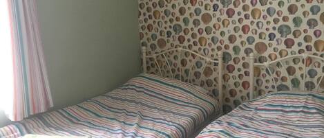 A room with two single beds, a patterned wallpaper, and a window with curtains.