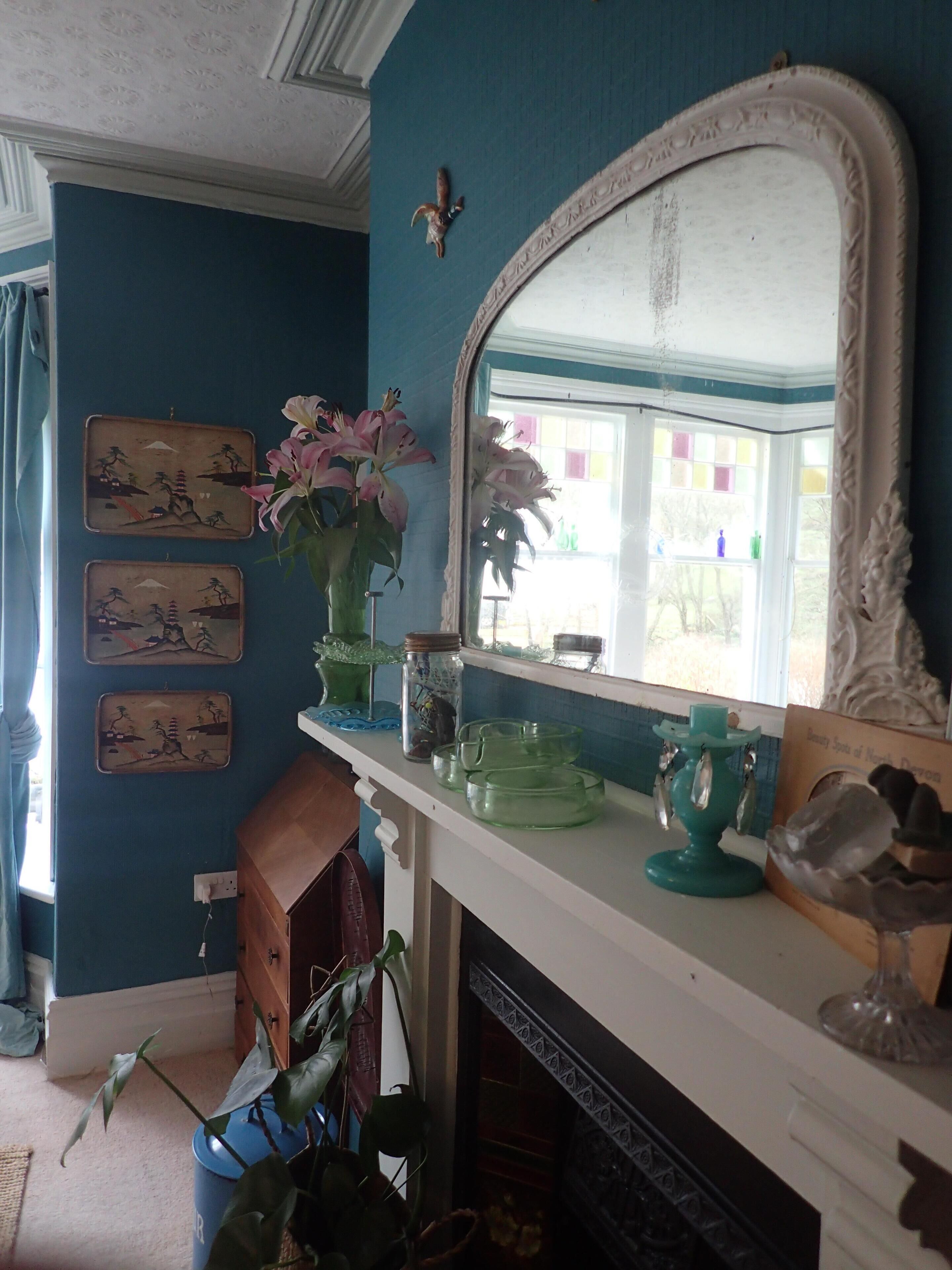 A room with a blue wall, a white fireplace, a mirror, and decorative items including a vase with flowers and a shelf with glassware.