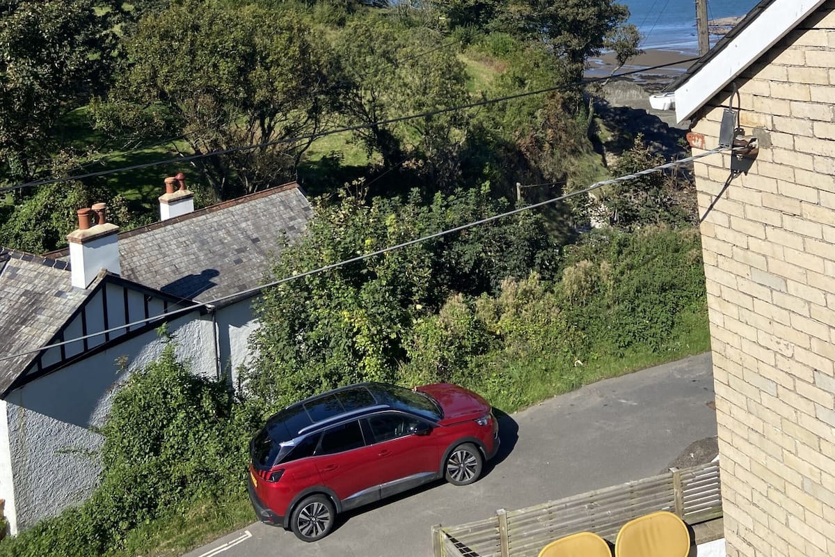 Une voiture rouge est garée dans une allée, offrant une vue sur un paysage côtier et une colline parsemée d’arbres.