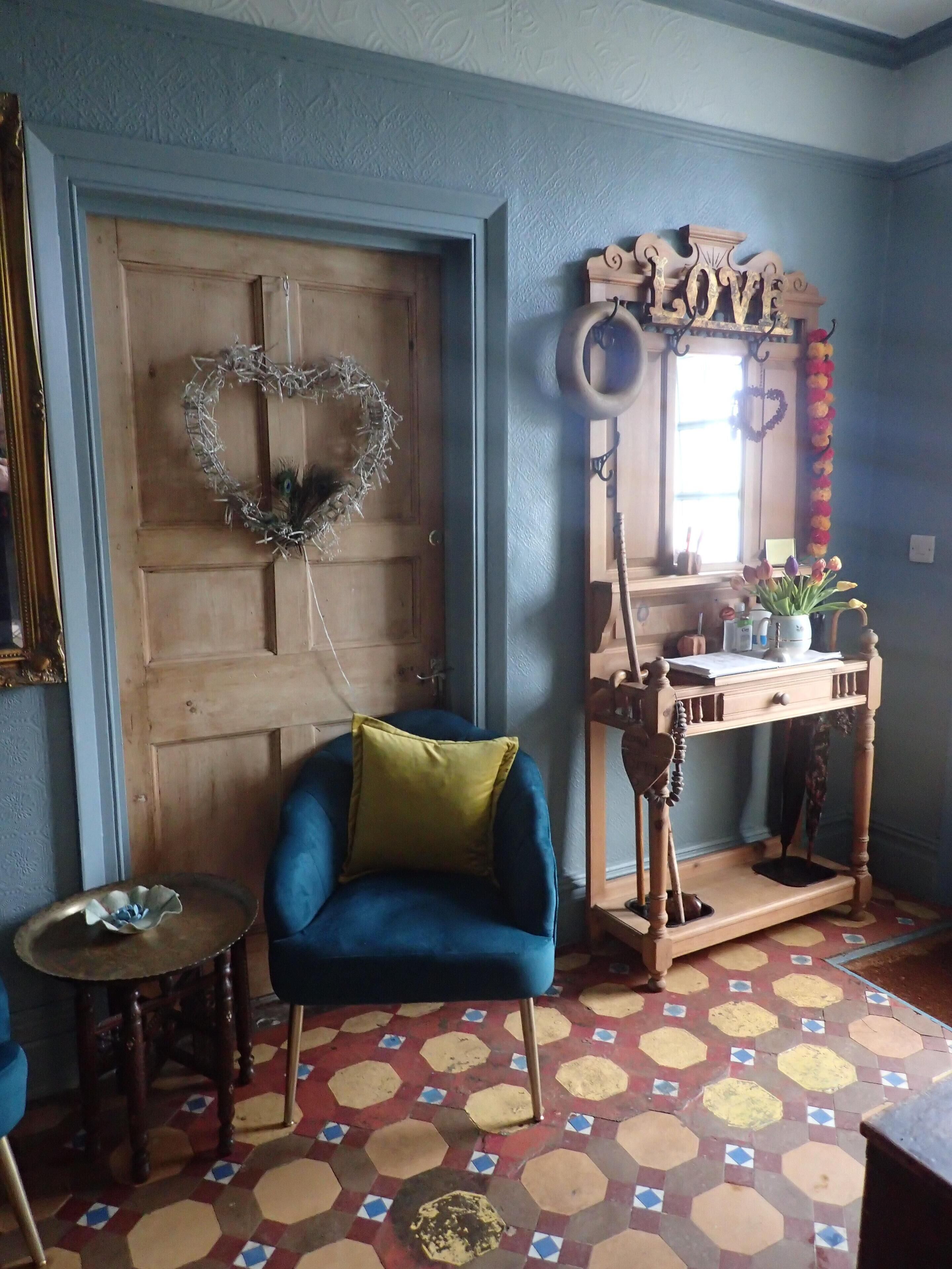A room with a wooden door, a blue armchair with a green cushion, a small round table, and a wooden console table with a mirror and decorative items.