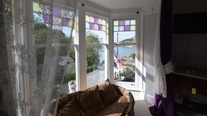 A room with a patterned sofa, a window with a view of a street and parked cars, and a curtain on the right. - Newberry Beach Lodge (Ilfracombe)