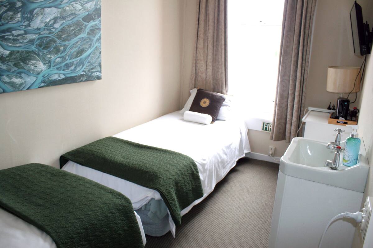 Une chambre d’hôtel avec deux lits simples, une salle de bain avec un lavabo et un miroir, et un tableau accroché au mur.