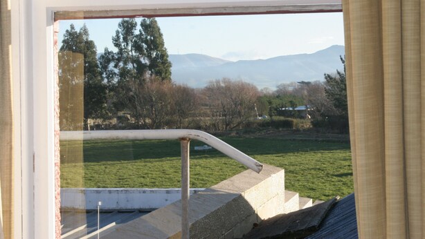 A view through a window with a metal railing, a grassy field, trees, and mountains in the distance. - Lumsden Hotel (Lumsden)