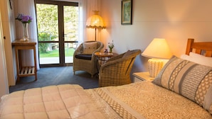 A hotel room with a bed, a wicker chair, a small table, a lamp, and a view of greenery outside. - Rolling Hills Country Stay B&B (Oropi)