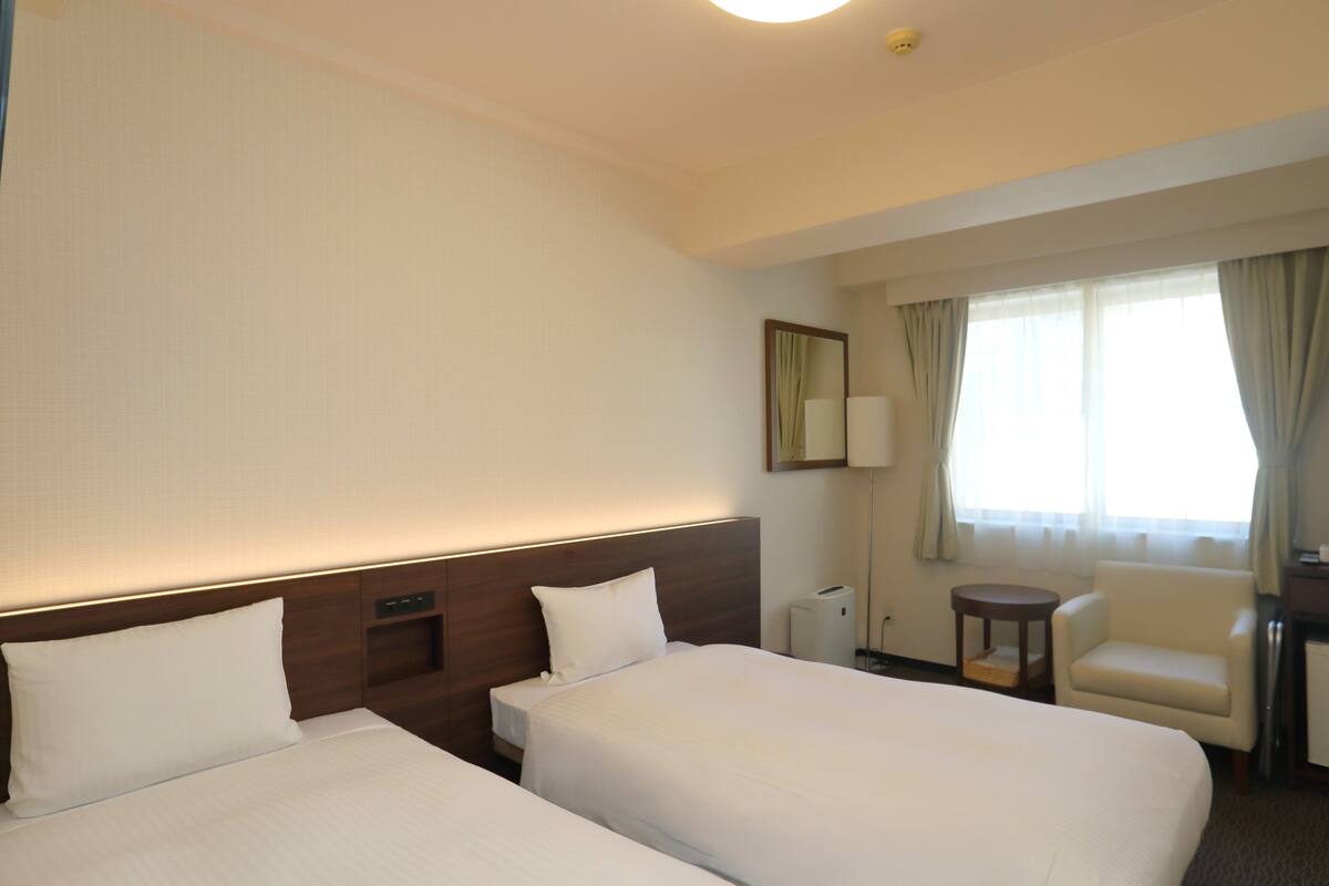 A hotel room with two beds, a chair, a desk, a lamp, and a window with curtains.