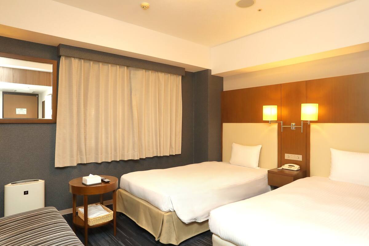 A hotel room with two beds, a bedside table with a lamp, a mirror, and a wall-mounted heater.
