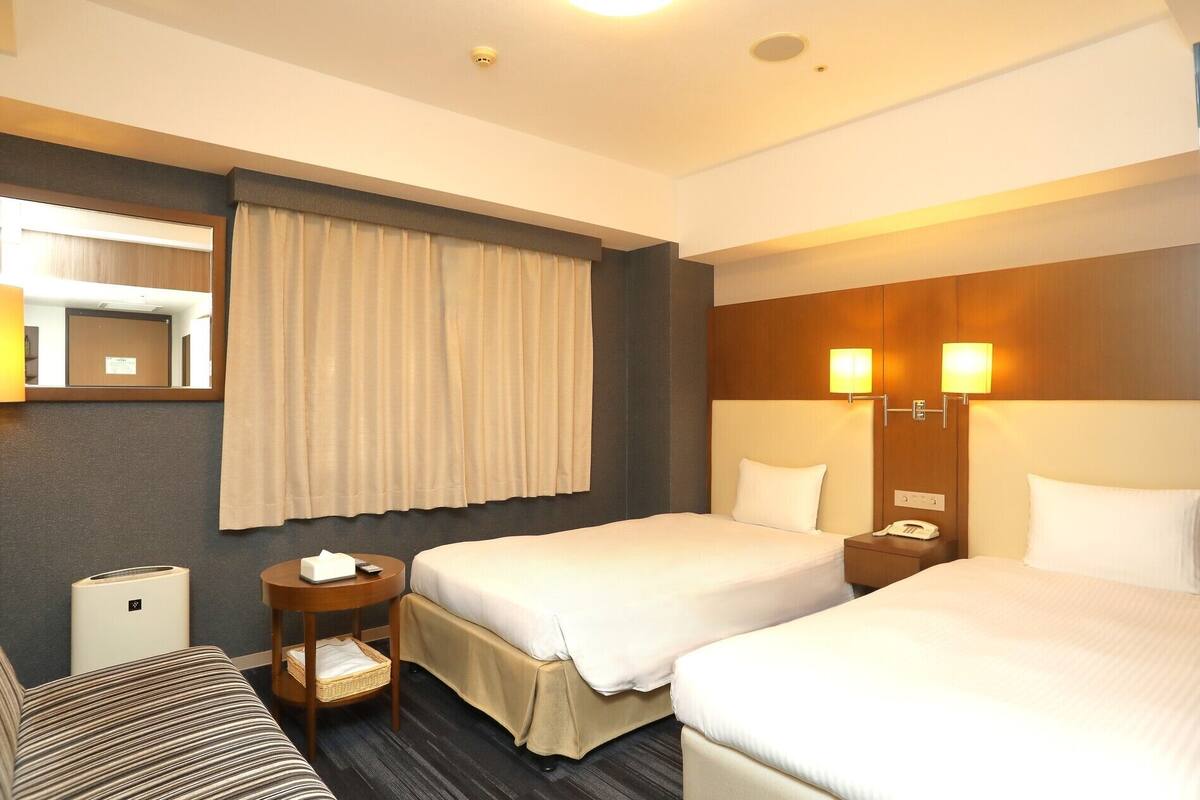 A hotel room with two beds, a bedside table with a lamp, a small table with a basket, a mirror, and a wall-mounted light fixture.