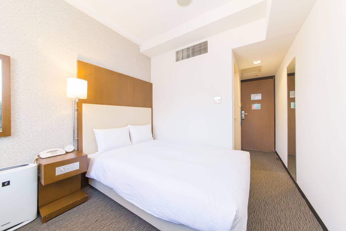 A hotel room with a large bed, a bedside table with a lamp, and a small heater.