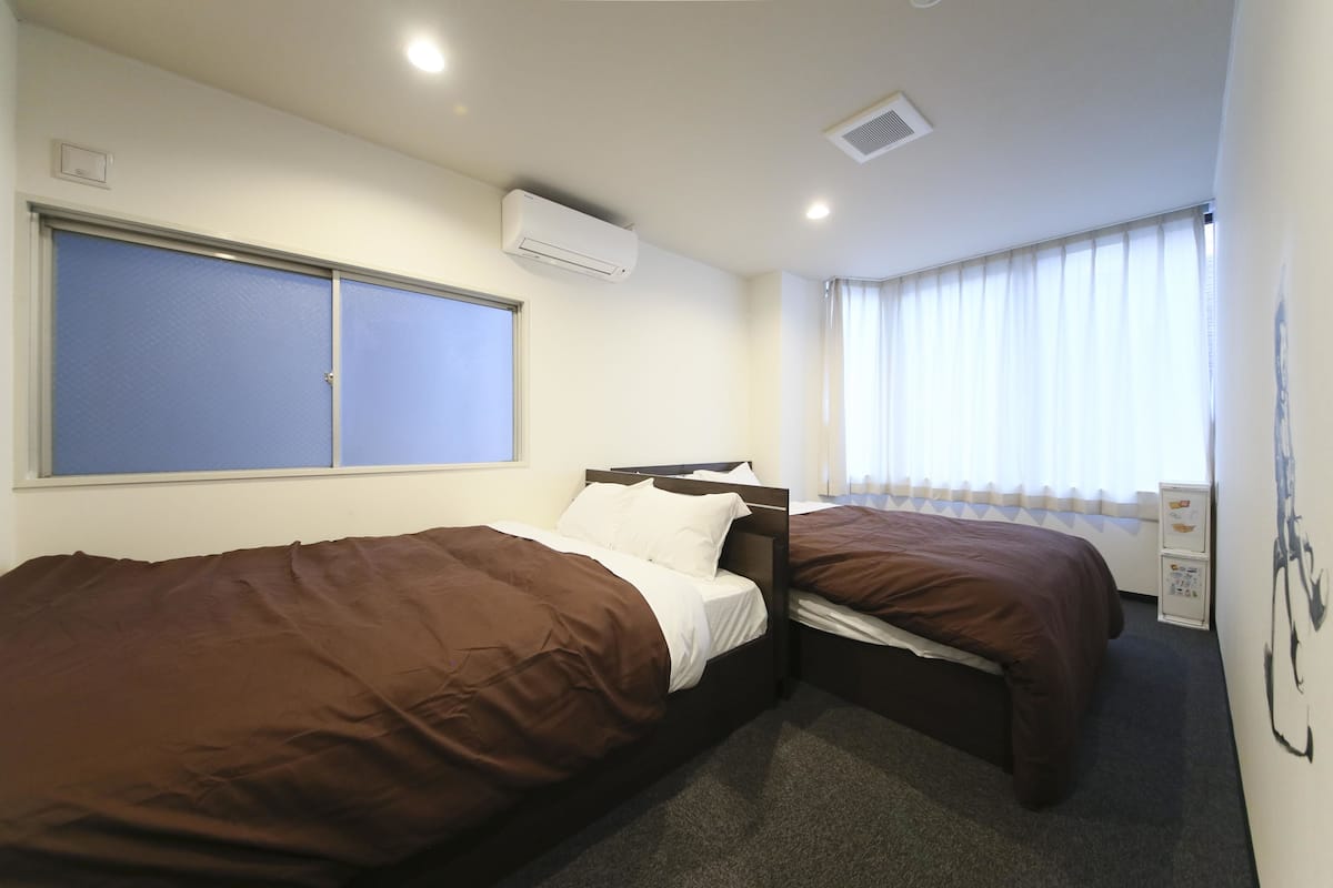 Habitación de hotel con dos camas individuales, una ventana con cortinas, un equipo de aire acondicionado y un armario pequeño con toma de corriente.