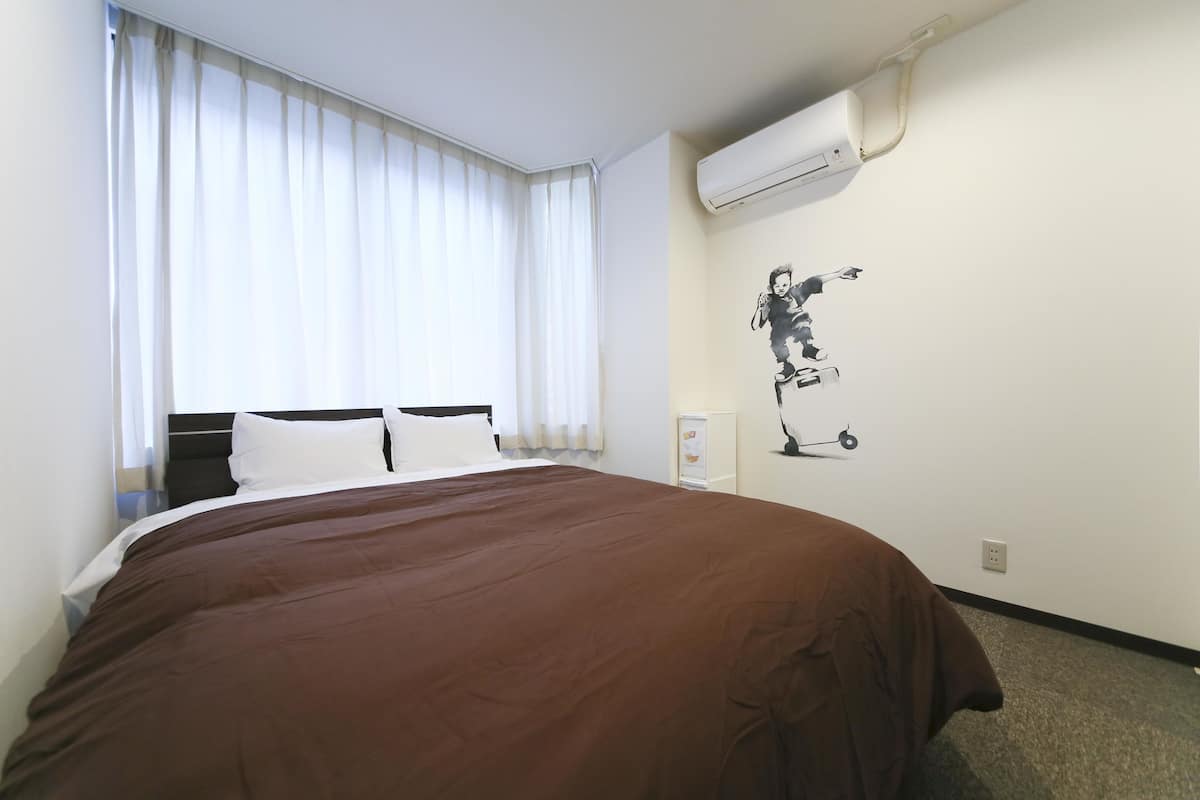 Habitación de hotel con una cama grande, una ventana con cortinas blancas, un equipo de aire acondicionado y un cuadro en la pared de una persona montando en bicicleta.