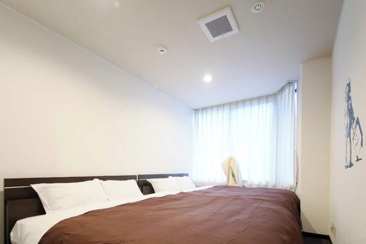 Una habitación de hotel con una cama grande, una ventana con cortinas, una obra de arte colgada en la pared y una rejilla de ventilación en el techo.