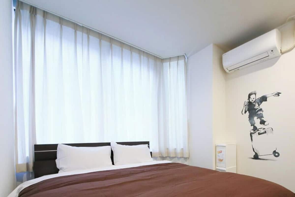 Habitación de hotel con cama, cortinas blancas, equipo de aire acondicionado y una obra de arte colgada en la pared.