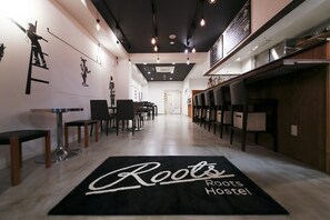 A hallway with a black carpet reading "Roots Hostel," wooden chairs, tables, and a bar area with stools. - Roots Hostel (Osaka)