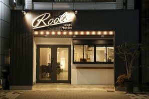 The exterior of the Roots Hostel with a modern design, featuring a prominent illuminated sign and a welcoming entrance.