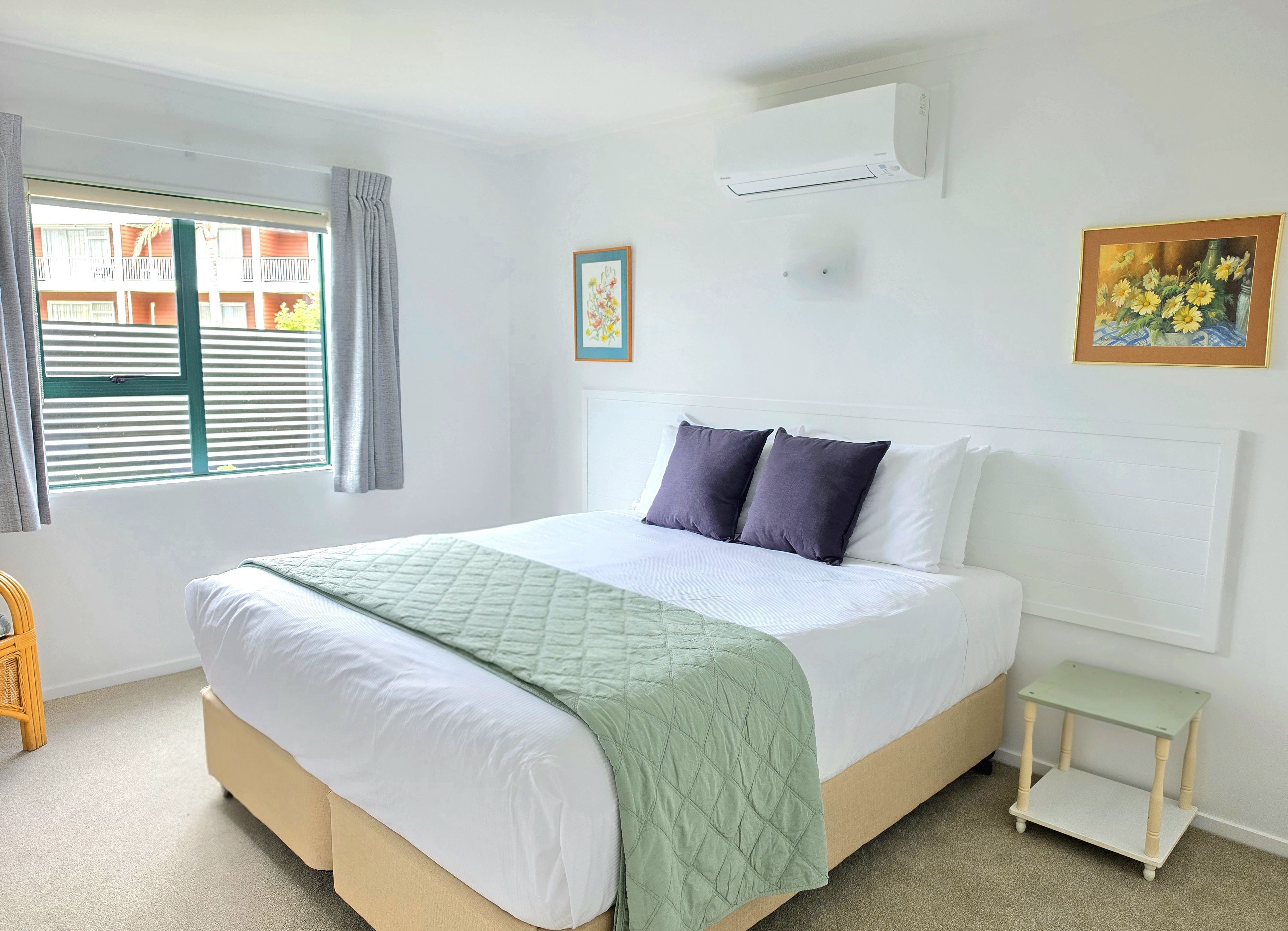 A bedroom with a bed, a window with blinds, an air conditioning unit, a bedside table, and a framed picture on the wall.