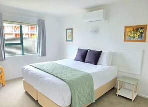 Een slaapkamer met een bed, een raam met jaloezieën, een airconditioning, een nachtkastje en een ingelijst schilderij aan de muur.