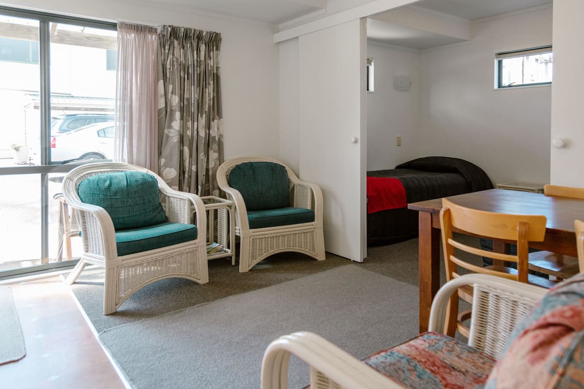 A room with a large window, two wicker chairs with green cushions, a wooden dining table, and a red blanket on a bed.