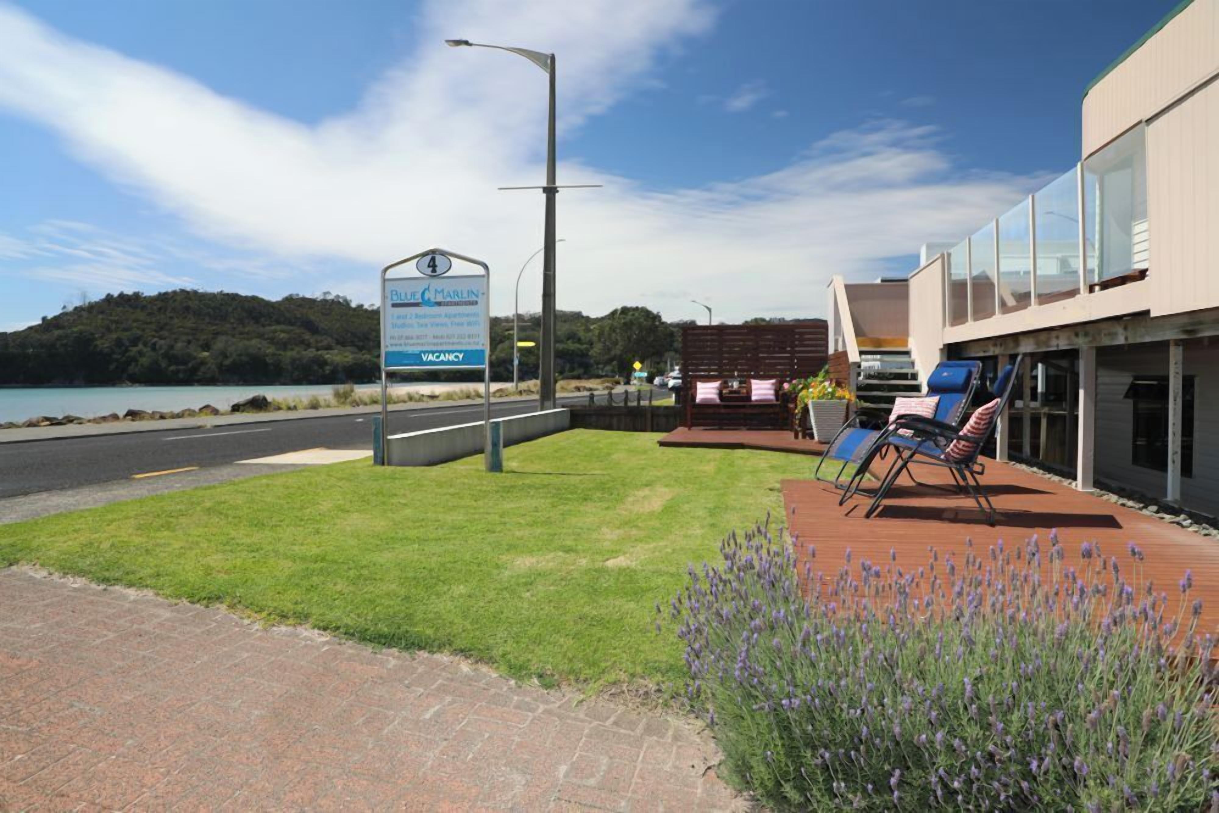 Un hôtel appelé Blue Marlin, avec une enseigne indiquant qu’il y a des chambres disponibles. Il dispose d’une zone pavée avec une pelouse, d’une terrasse en bois avec des chaises longues et offre une vue sur la mer.