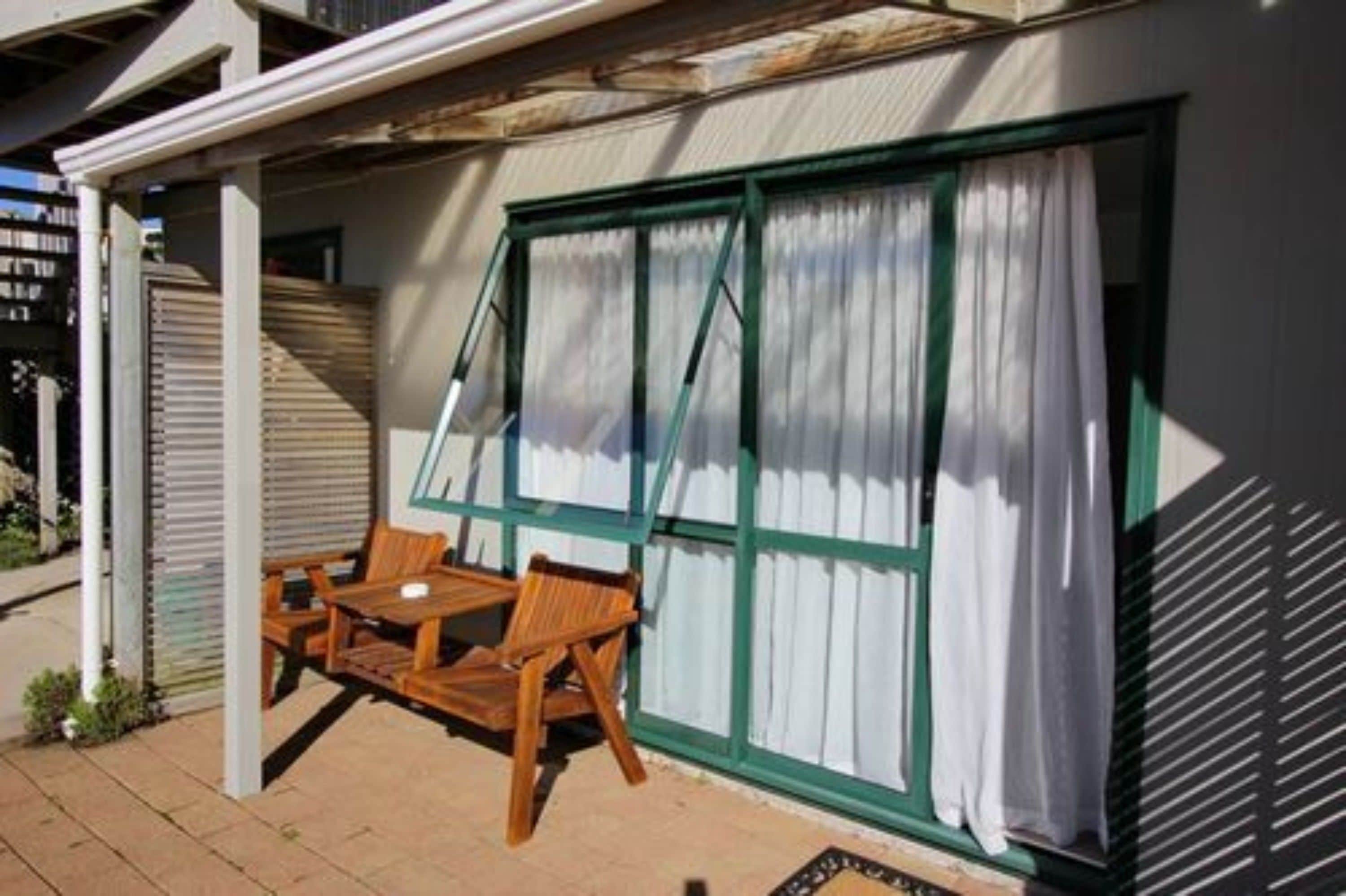 A covered patio with a wooden table and chairs, a window with green frames and white curtains, and a metal shutter door.