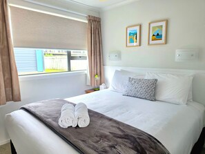 Een hotelkamer met een bed, twee kussens, een dekbed, een raam met gordijnen en twee foto's in lijsten aan de muur.