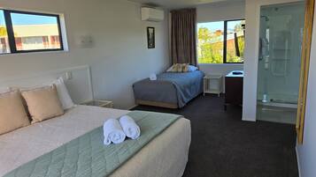 A hotel room with two beds, a small table, a shower area, and a window with a view of buildings and trees.