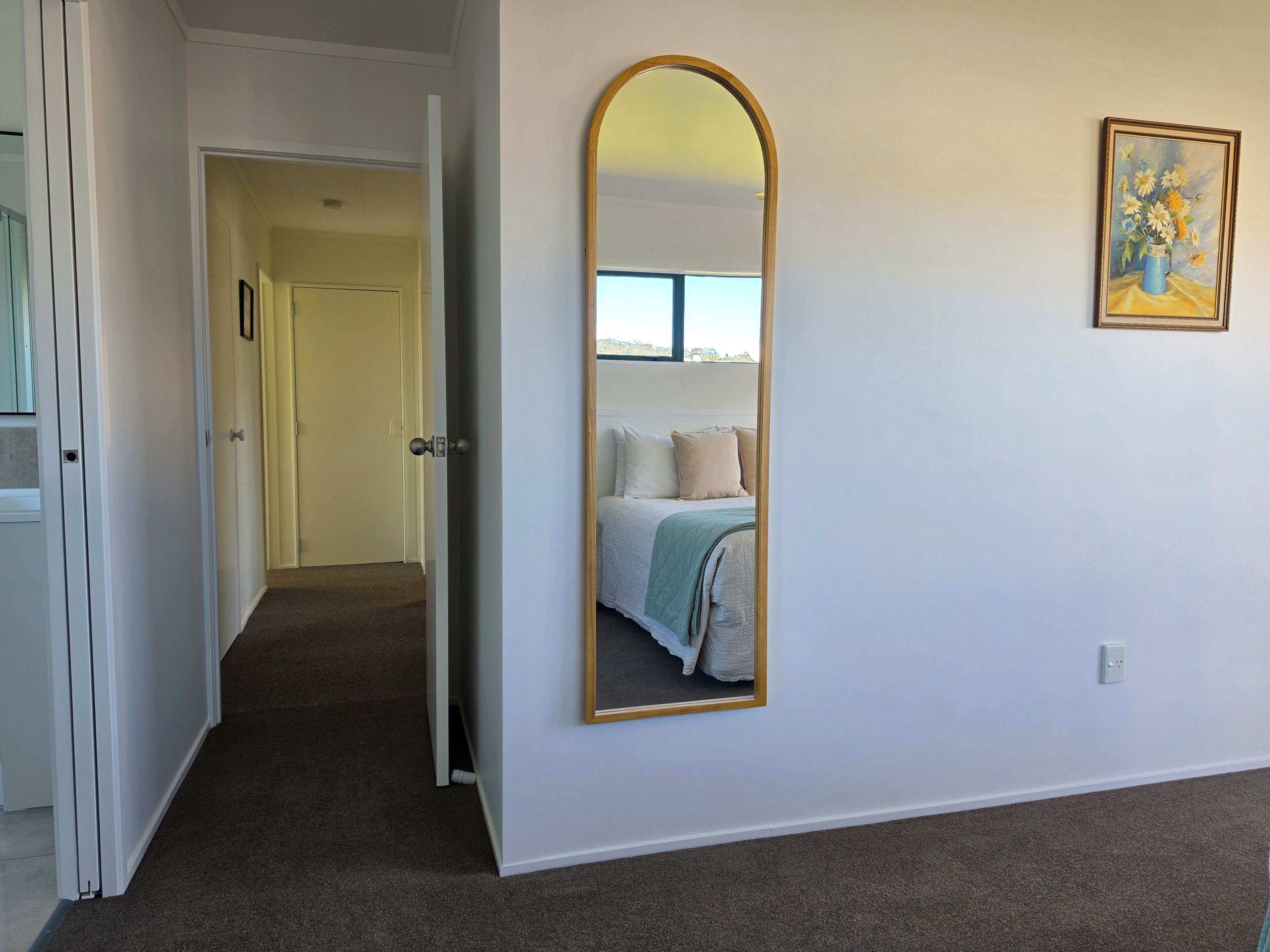 Un dormitorio con un espejo grande, una cama con almohadas, una puerta que da a otra habitación y un cuadro en la pared.