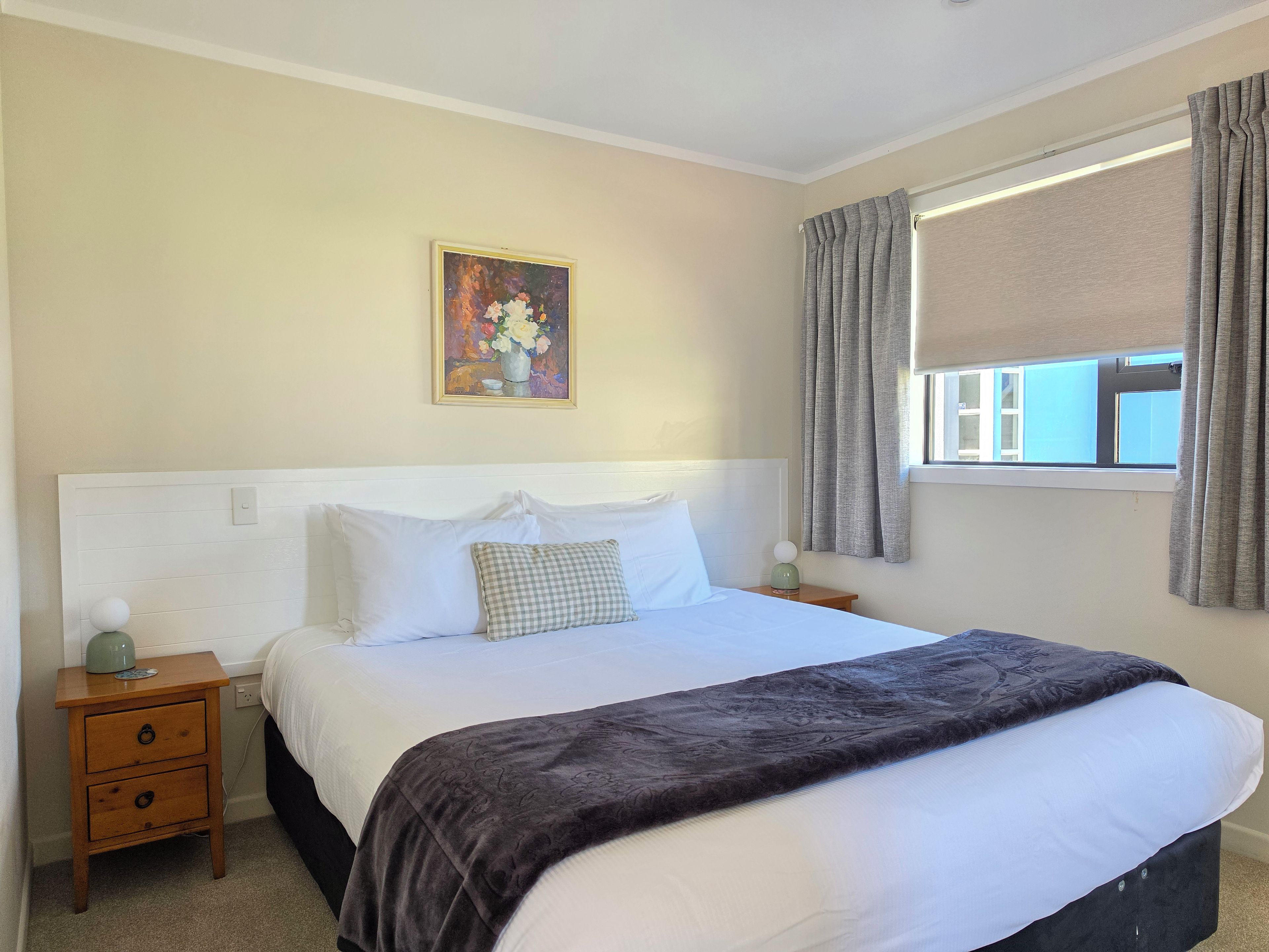 A bedroom with a bed, a nightstand, a painting, a window with curtains, and a view of the outside.