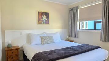 A bedroom with a bed, a nightstand, a painting, a window with curtains, and a view of the outside.