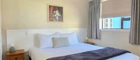 A bedroom with a bed, a nightstand, a painting, a window with curtains, and a view of the outside.