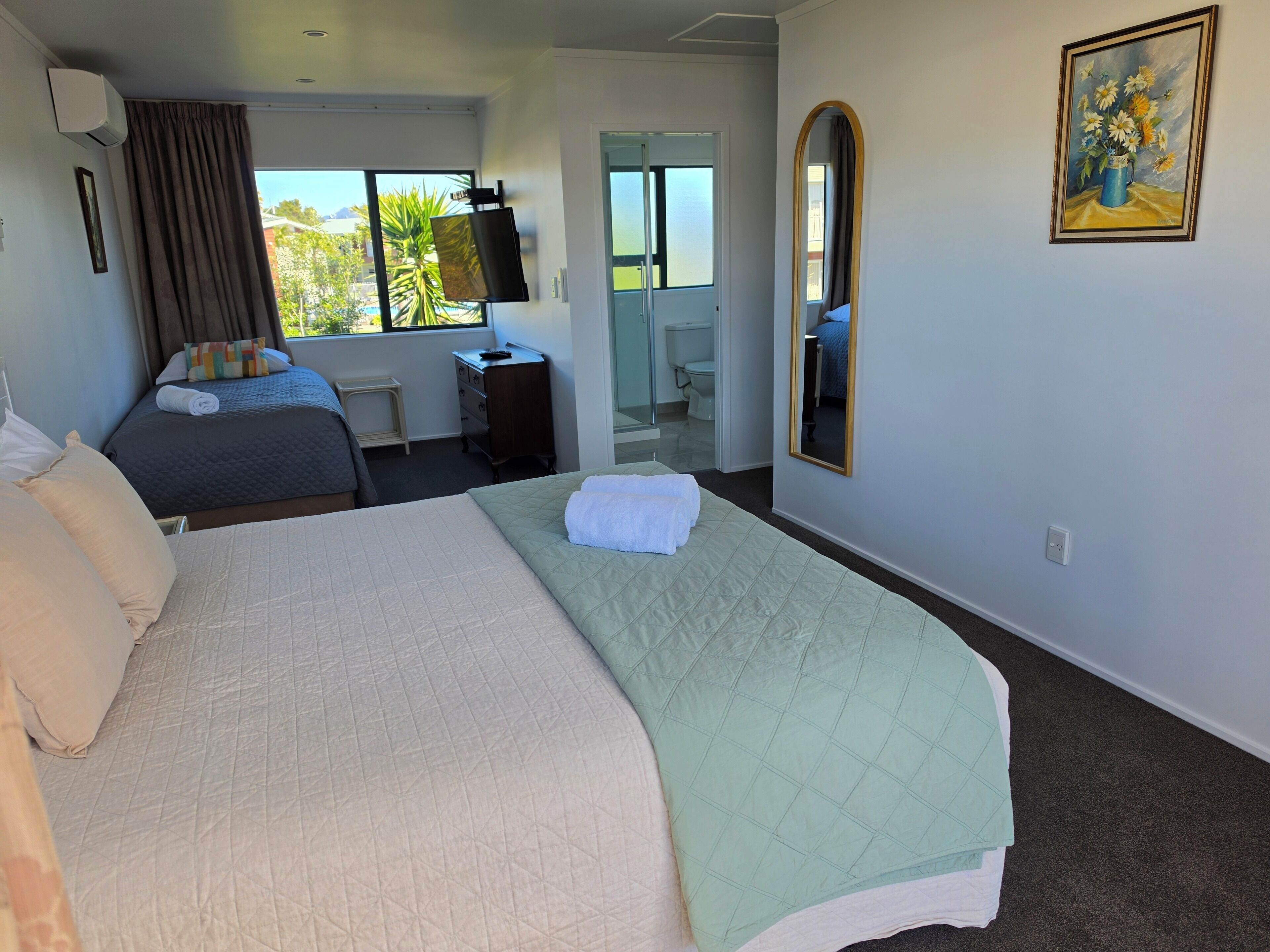 Ein Hotelzimmer mit einem großen Bett, einem Schreibtisch, einem Spiegel und einem Fenster mit Blick auf Grünflächen.