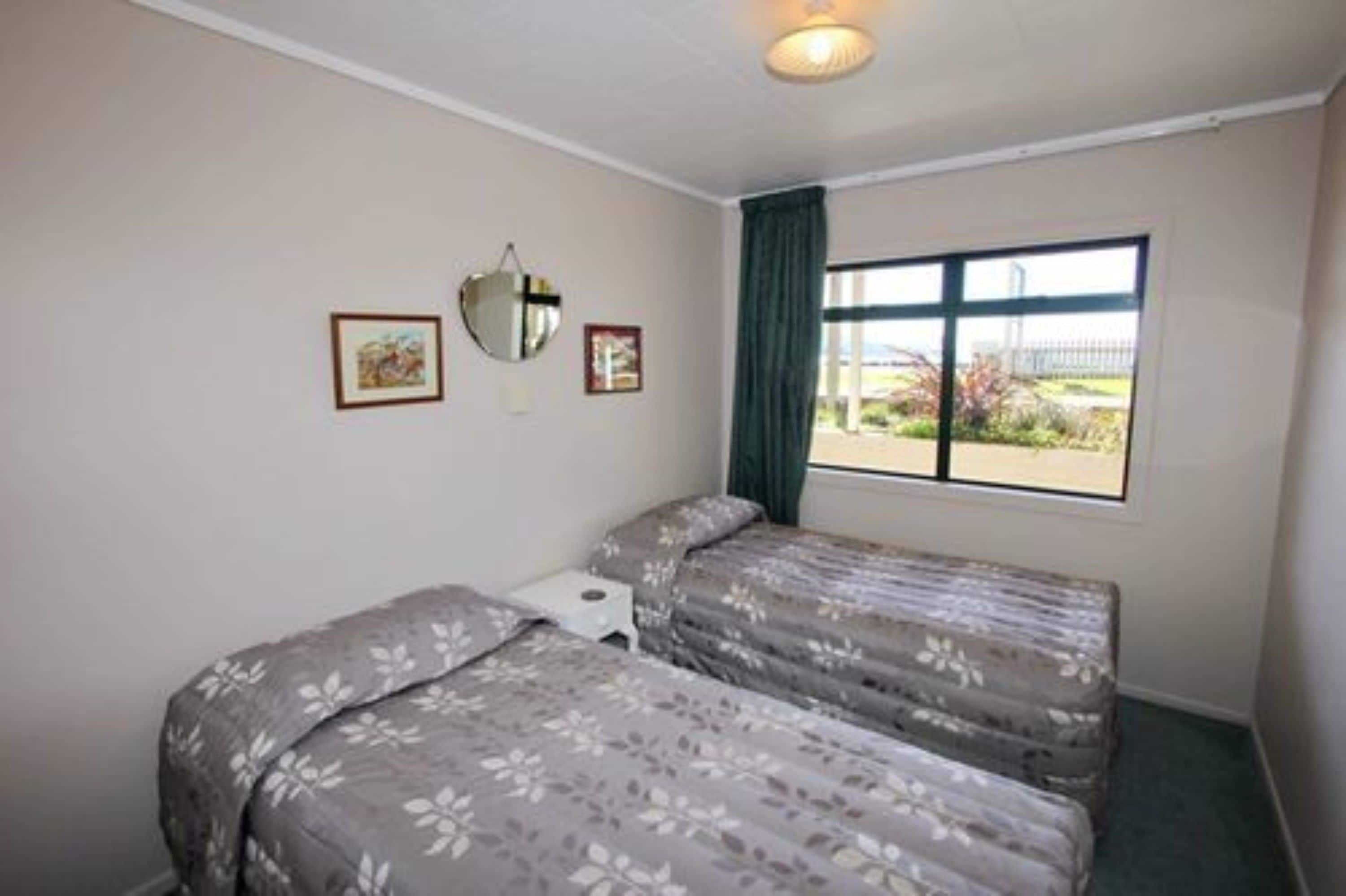 A room with two beds, a small bedside table, a window with a view of the outdoors, and a ceiling light.