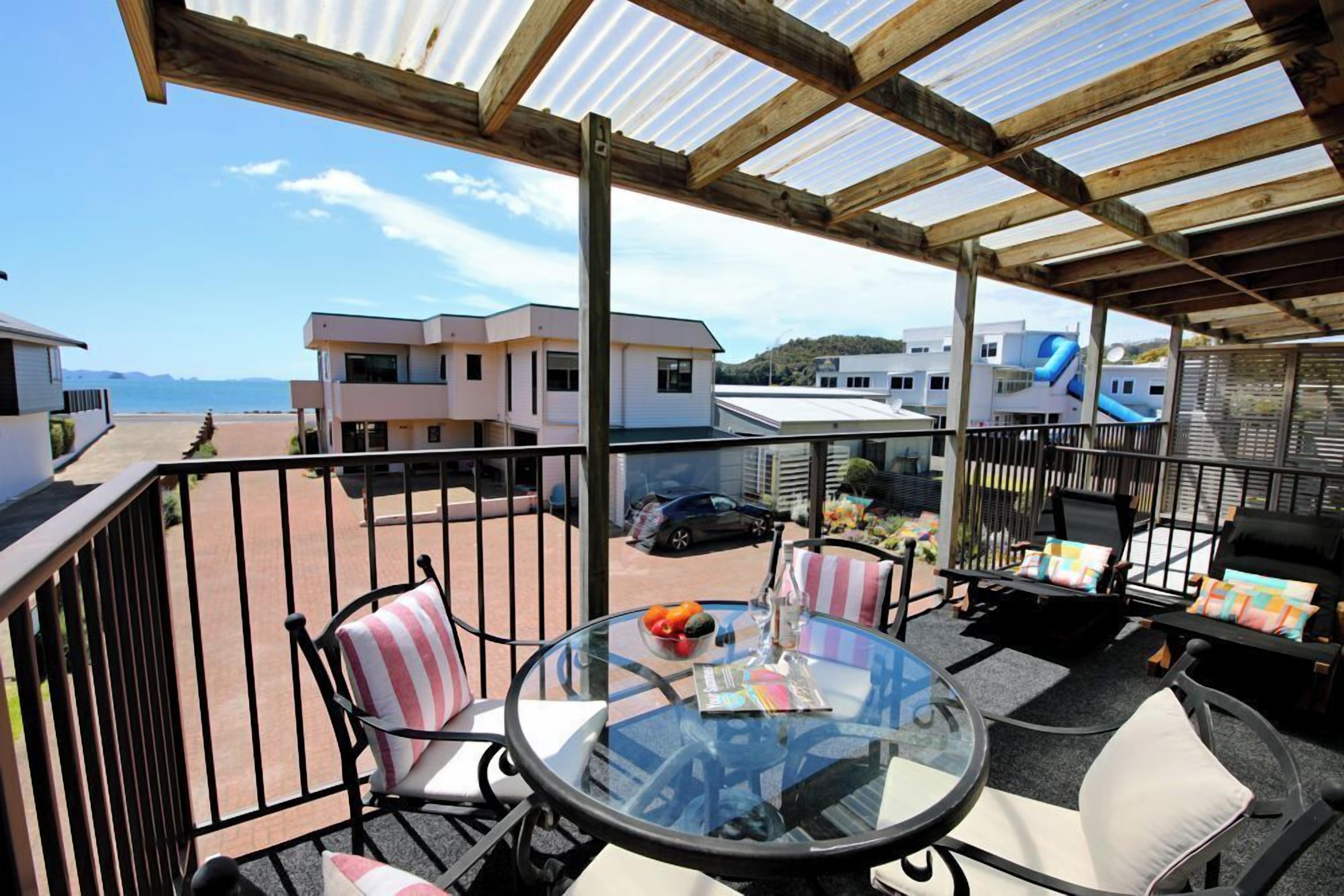 A covered outdoor seating area with a glass table, chairs with striped cushions, and a view of buildings and a car in the background.