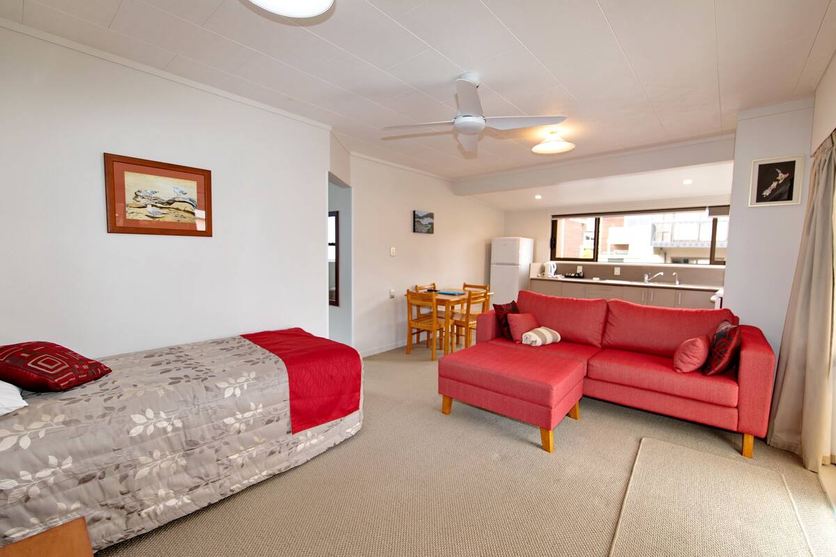 A hotel room with a bed, a red sofa, a dining table, and a kitchen area.