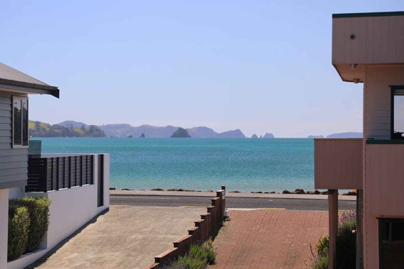 Une propriété située sur la côte, avec un chemin pavé menant à la plage, un bâtiment moderne doté d’un balcon et une vue dégagée sur la mer et les îles lointaines.