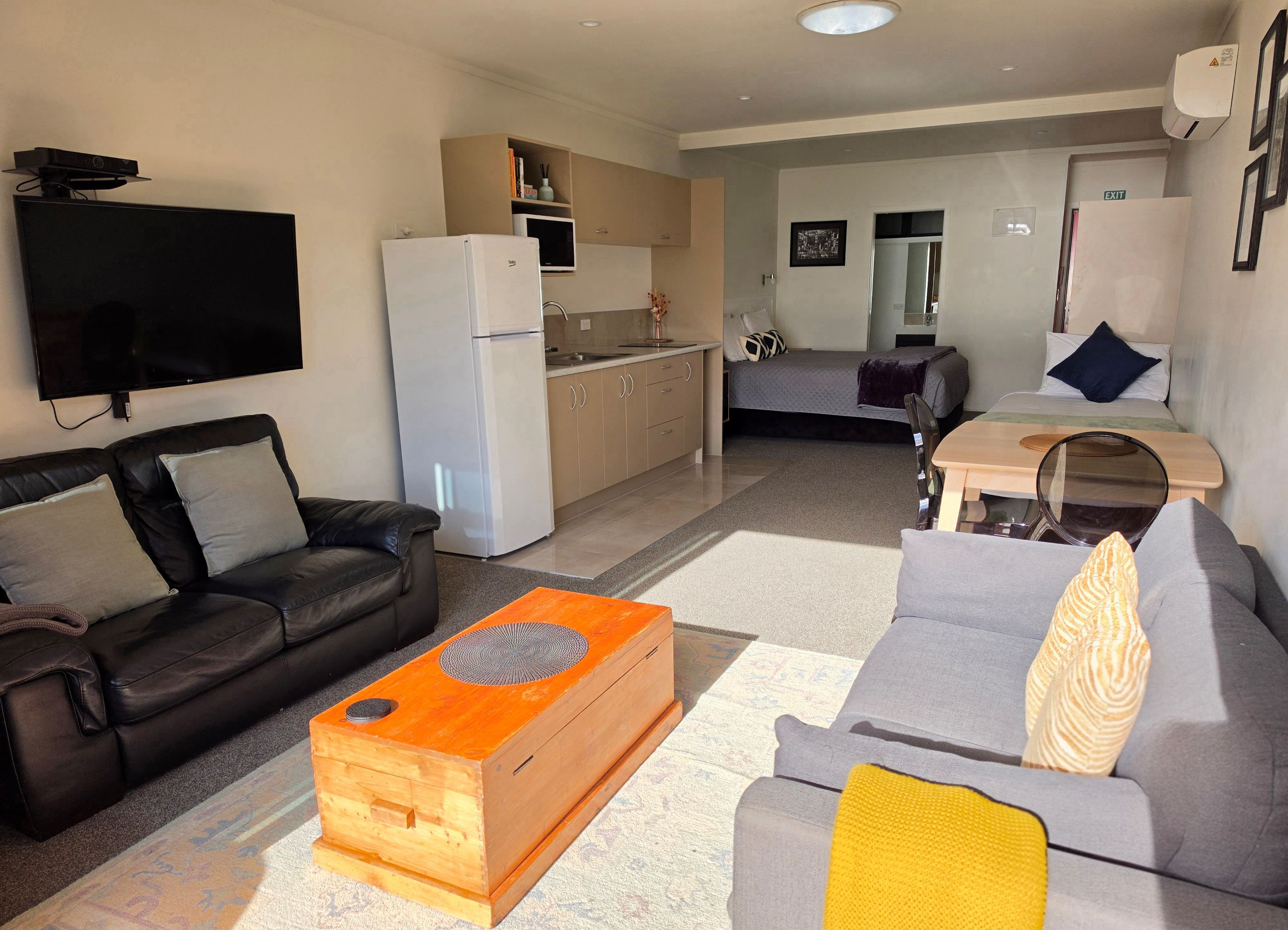 A modern living room with a flat-screen TV, a black leather sofa, a grey sofa, a wooden coffee table, and a kitchen area with a refrigerator and microwave.