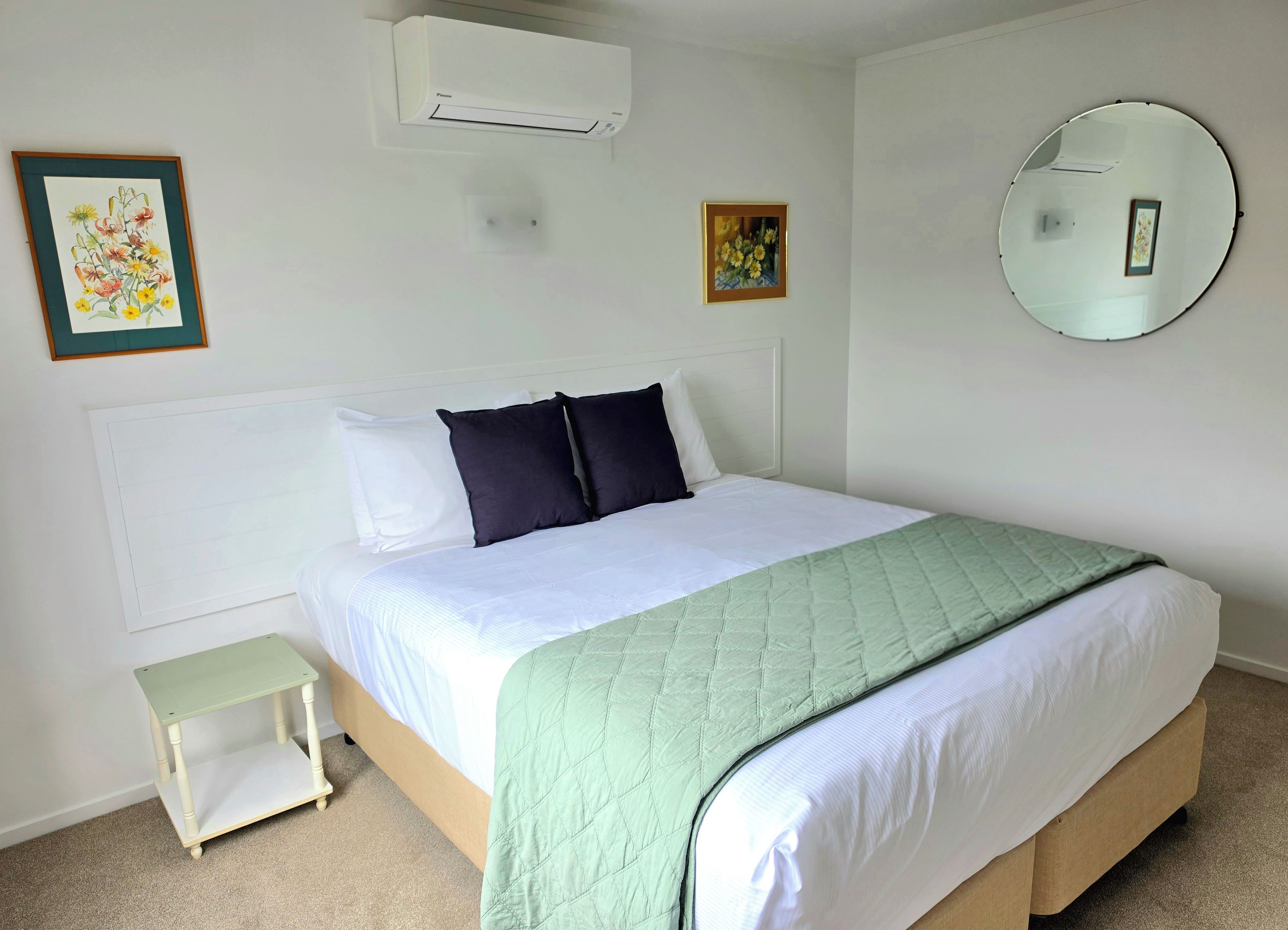 Una cama bien hecha con colcha blanca y verde, una mesita de noche pequeña y un aire acondicionado de pared.