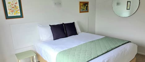 Una cama bien hecha con colcha blanca y verde, una mesita de noche pequeña y un aire acondicionado de pared.
