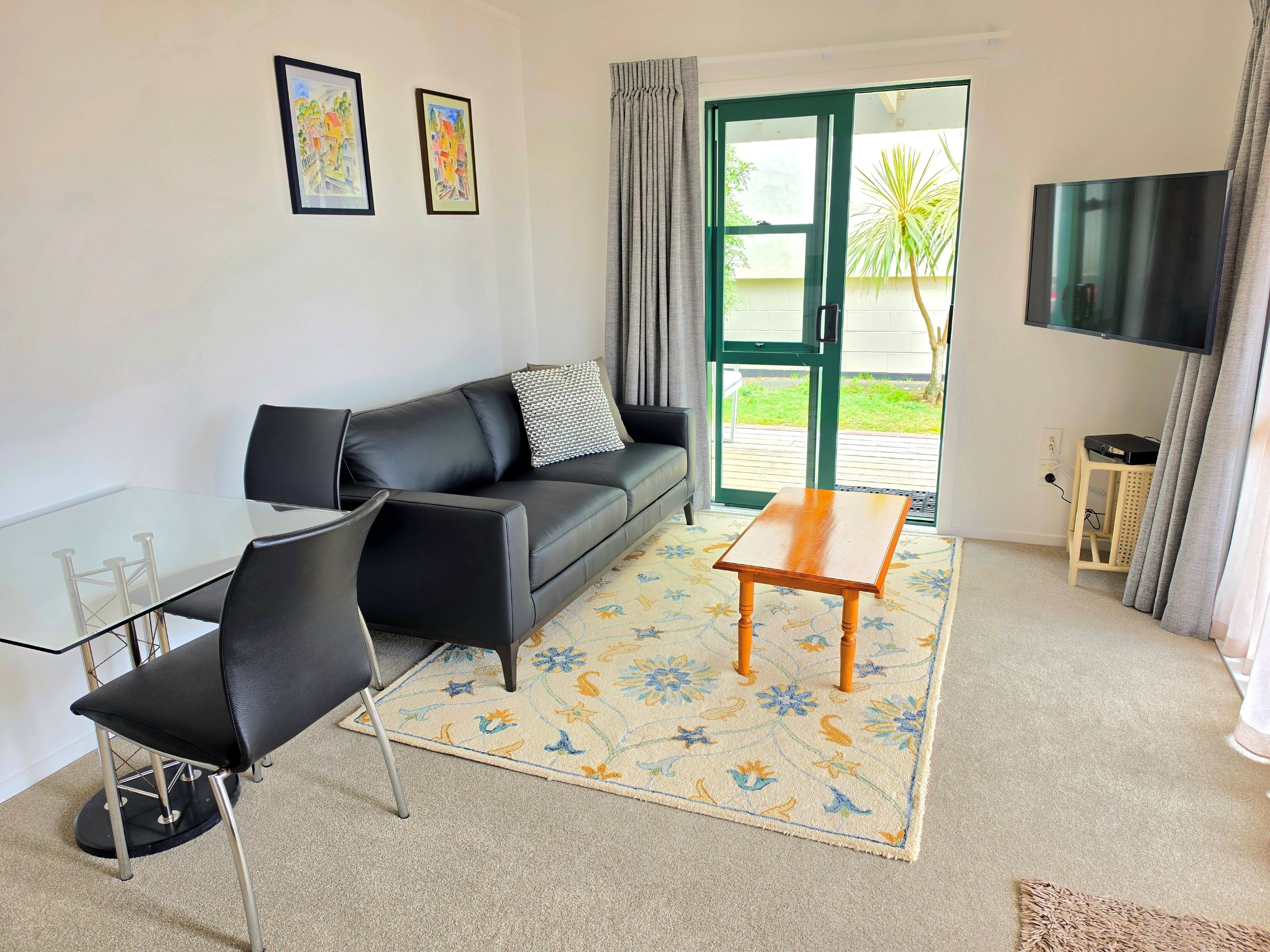 A living room with a glass table, black sofa, and a wooden coffee table. There is a TV mounted on the wall and a window with curtains.