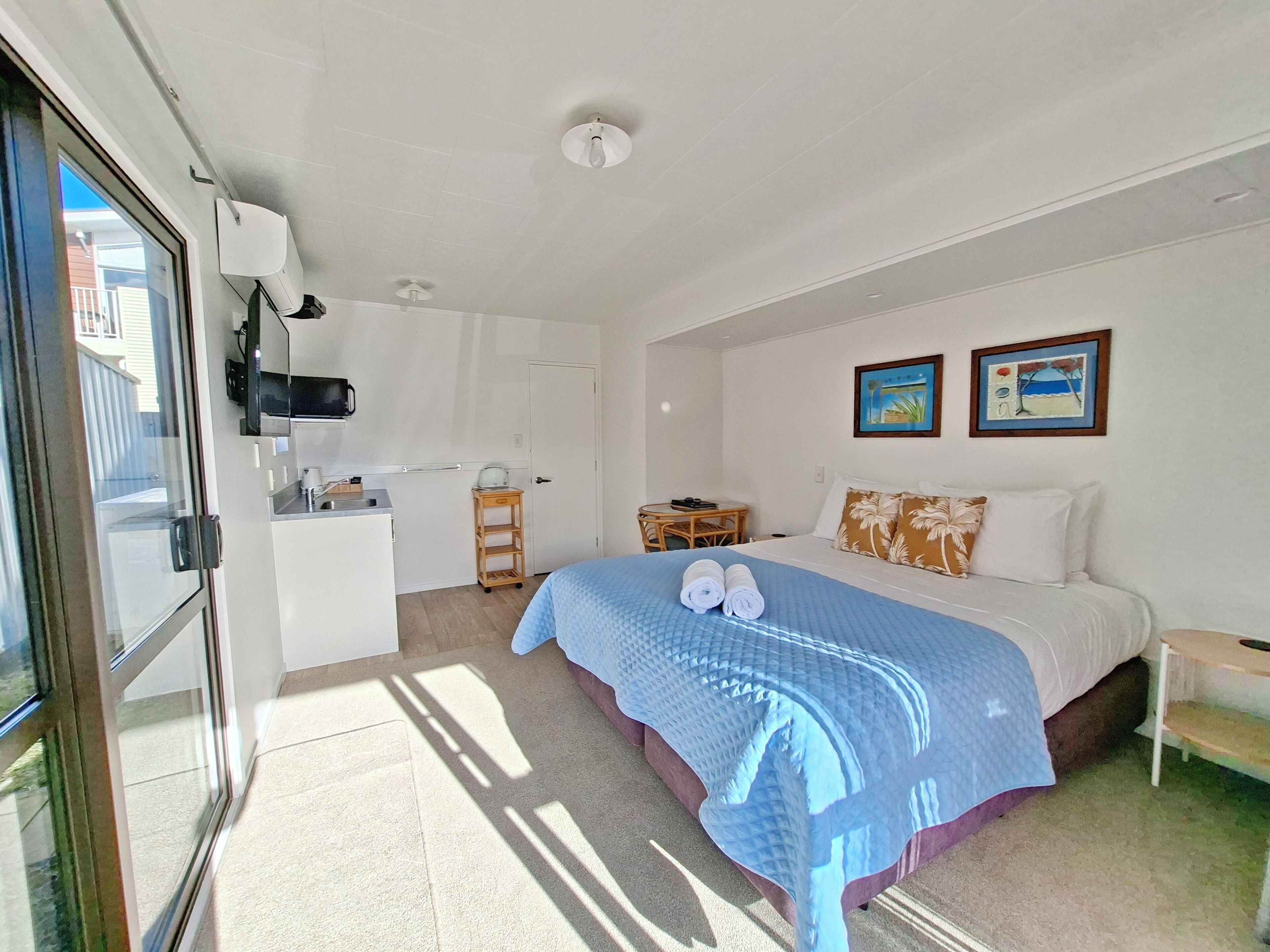 A hotel room with a bed, a small table, a chair, a kitchenette, and a door leading outside.